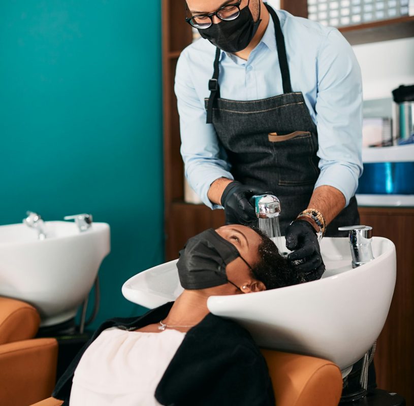 Hairdresser wearing face mask while washing black woman's hair at the salon.